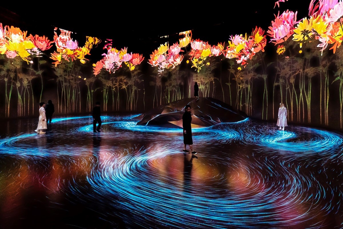 Instalação artística no museu teamLab, telas gigantes exibem flores enquanto pessoas caminham sobre um projeção, onde seus rastros de linhas azuis seguem os seus passos.